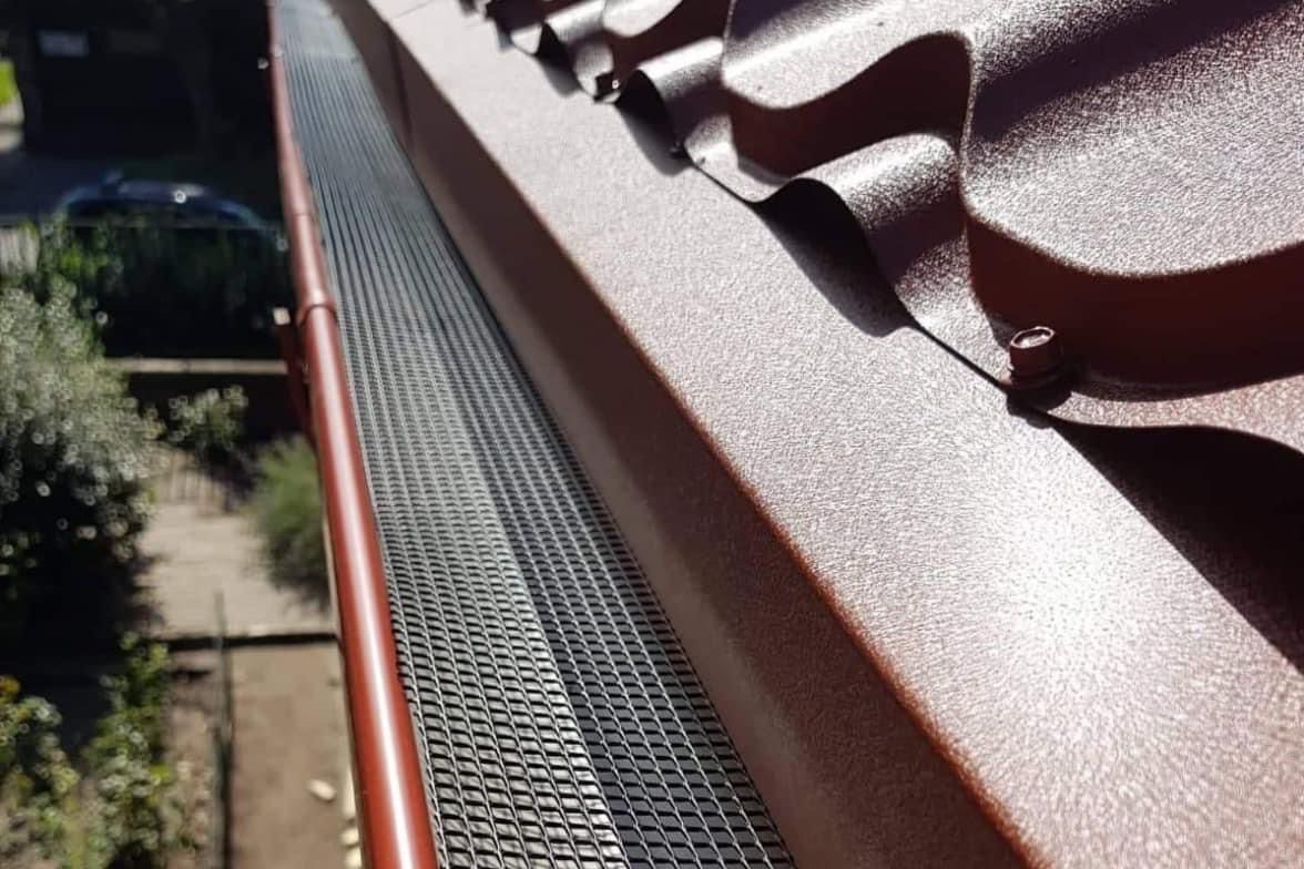 Brązowy dach z metalową siatką zabezpieczającą rynnę przed liśćmi. Widok z góry, ostre światło słoneczne podkreśla fakturę dachówki i siatki.