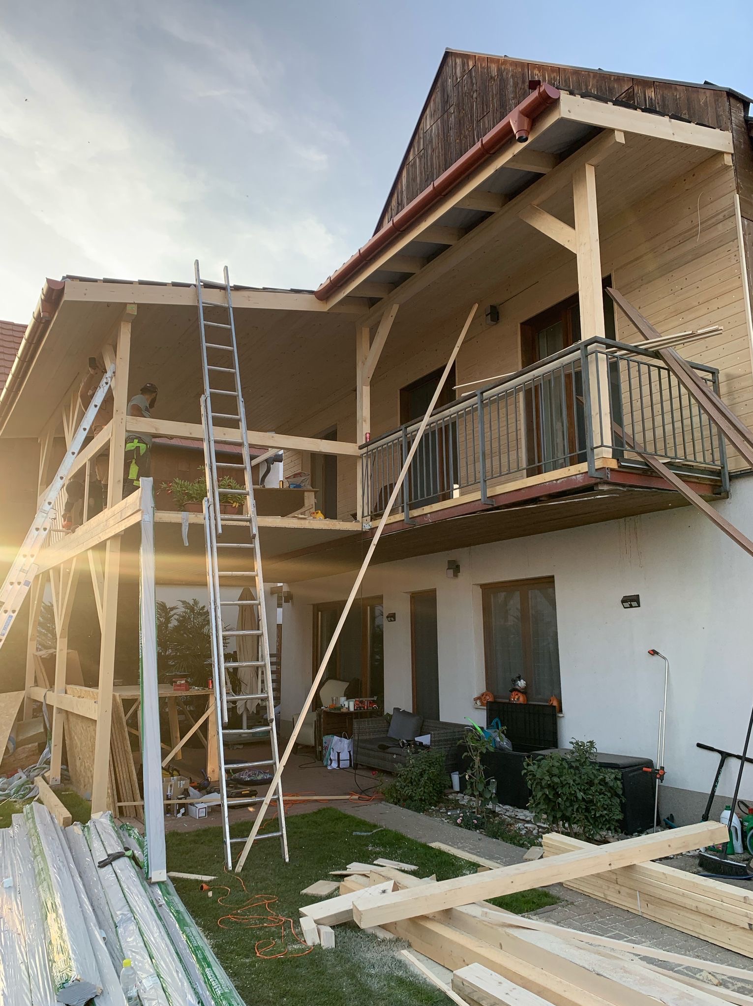 Konstrukcja drewniana domu w trakcie budowy, widoczne rusztowania i drabiny. Częściowo zamontowane rynny. W tle jasne słońce.