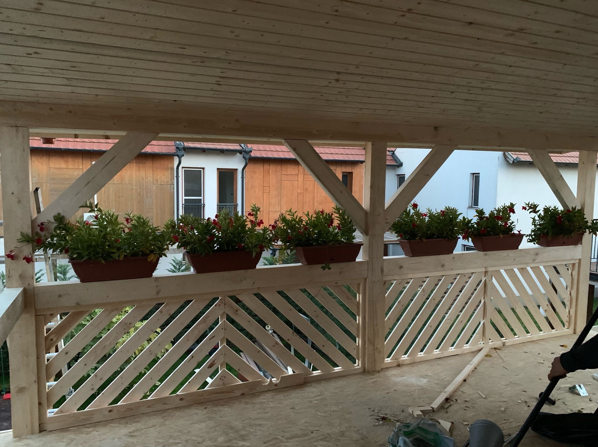 Drewniana konstrukcja z ukośnymi listwami i donicami z kwiatami na balkonie, widok na budynki w tle. Elementy konstrukcyjne z surowego drewna.