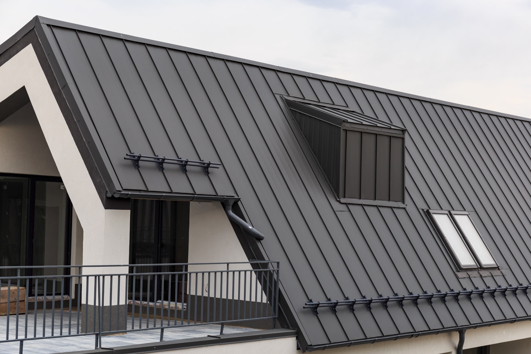 Nowoczesny dom z grafitowym dachem z blachy na rąbek stojący, okna dachowe i orynnowanie w tym samym kolorze. Widok z dołu, fragment balkonu z szarą balustradą.