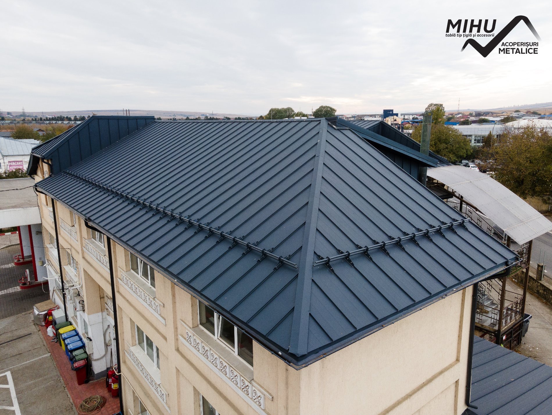 Nowoczesny, ciemnoszary dach z blachy trapezowej na budynku z beżową elewacją, widok z góry. W tle zabudowa miejska i pochmurne niebo. Widoczny logotyp firmy.