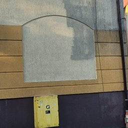 USŁUGI DŹWIGOWE ROBERT CIEŚLA - Częściowo umyta elewacja budynku z żółtą skrzynką gazową i wiadrem na chodniku. Widoczny podnośnik koszowy. Ujęcie z dołu, widać różnicę w czystości.