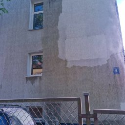 USŁUGI DŹWIGOWE ROBERT CIEŚLA - Fragment elewacji budynku po częściowym umyciu, widoczne różnice w kolorze. Dwa okna, siatka ogrodzeniowa i zaparkowany samochód w tle. Tabliczka z numerem budynku.