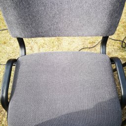 USŁUGI DŹWIGOWE ROBERT CIEŚLA - Szare krzesło biurowe z czarnymi podłokietnikami, ustawione na trawie. Widoczny fragment białego buta w dolnym rogu. Ujęcie z góry.