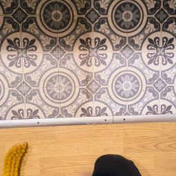 USŁUGI DŹWIGOWE ROBERT CIEŚLA - Podłoga po umyciu: wzorzyste płytki ceramiczne są czyste, a drewniana podłoga lśni. Obok leży żółta szczotka i widać fragment spodni osoby sprzątającej.