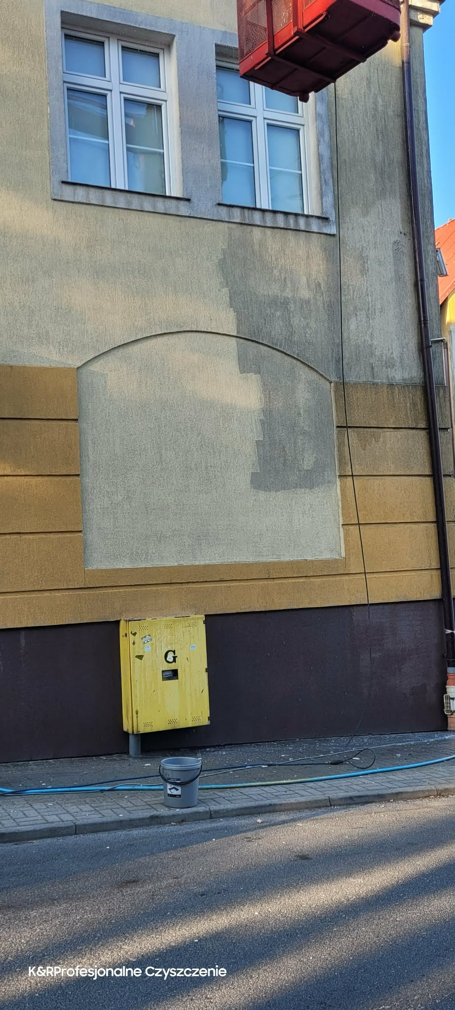 Częściowo umyta elewacja budynku z żółtą skrzynką gazową i wiadrem na chodniku. Widoczny podnośnik koszowy. Ujęcie z dołu, widać różnicę w czystości.