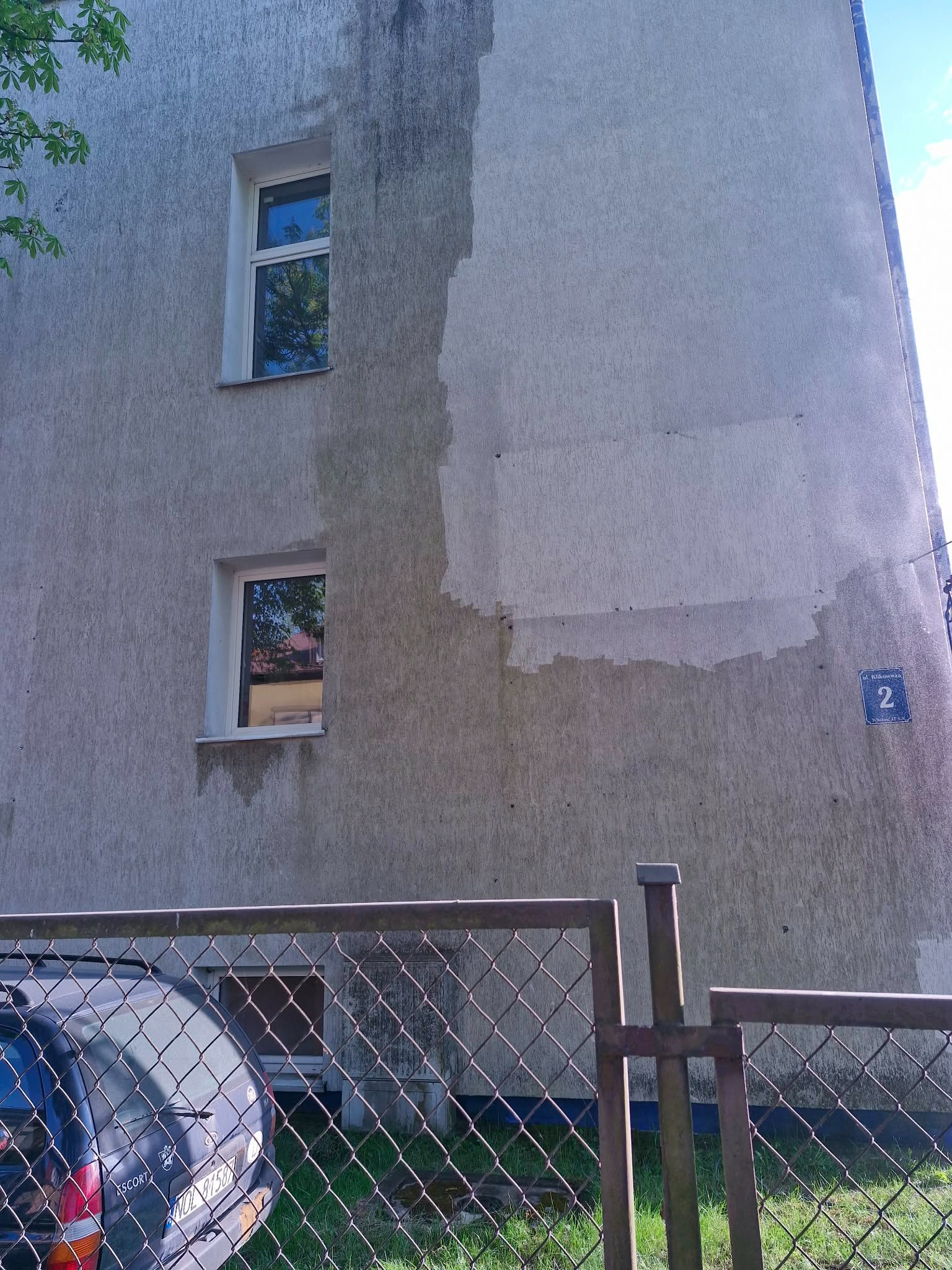 Fragment elewacji budynku po częściowym umyciu, widoczne różnice w kolorze. Dwa okna, siatka ogrodzeniowa i zaparkowany samochód w tle. Tabliczka z numerem budynku.