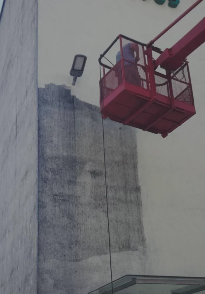 Pracownik na podnośniku czyści elewację budynku. Widoczny fragment wyczyszczonej ściany, lampa uliczna i fragment szklanego zadaszenia.