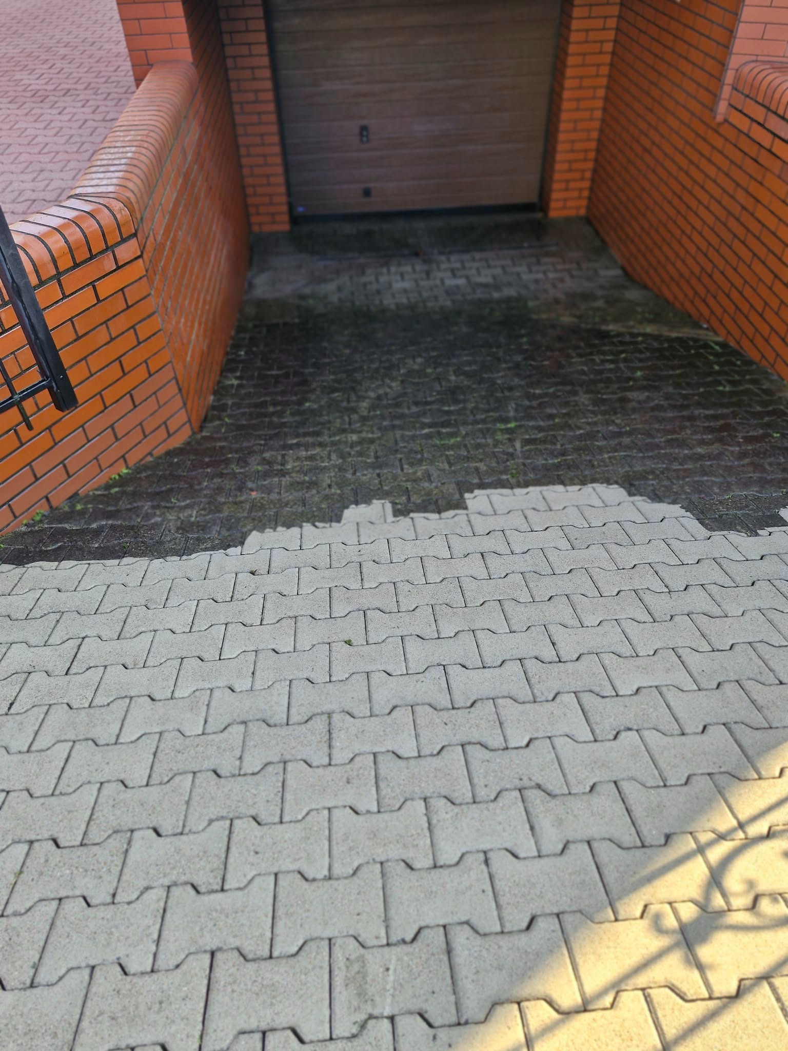Częściowo umyta kostka brukowa przed garażem, widoczna różnica w kolorze i czystości. Ceglana ściana po bokach, brama garażowa w tle. Ujęcie z góry.