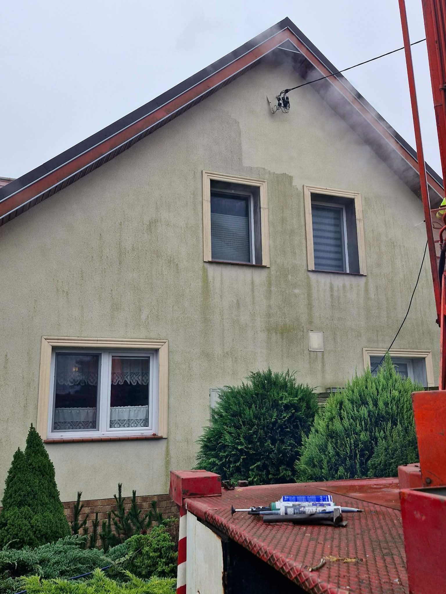 Czyszczenie elewacji domu z zielonego nalotu za pomocą myjki ciśnieniowej z podnośnika. Widoczne narzędzia na platformie. Częściowo umyta ściana budynku.