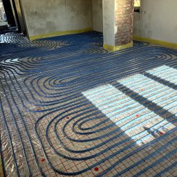 Jakub Błażejak - Niebieskie rury ogrzewania podłogowego ułożone na folii z czerwoną siatką, widoczne fragmenty ścian w surowym stanie i kolumna z cegły. Żółta taśma zabezpieczająca.