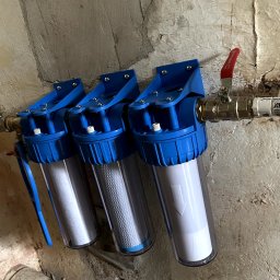 Jakub Błażejak - Potrójny filtr wody z niebieskimi obudowami zamontowany na ścianie z widocznymi rurami i zaworami. Filtracja wody w instalacji domowej.