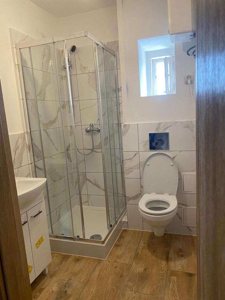 Wykończona łazienka z kabiną prysznicową, umywalką z szafką i toaletą. Płytki imitujące marmur na ścianach, drewnopodobne panele na podłodze. Małe okno w tle.