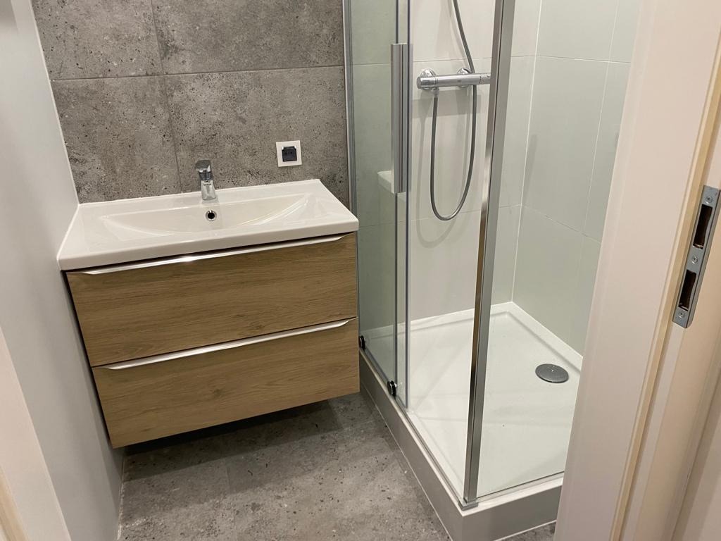 Nowoczesna łazienka z drewnianą szafką pod umywalką i kabiną prysznicową ze srebrnymi profilami na tle szarych płytek. Minimalistyczny design.