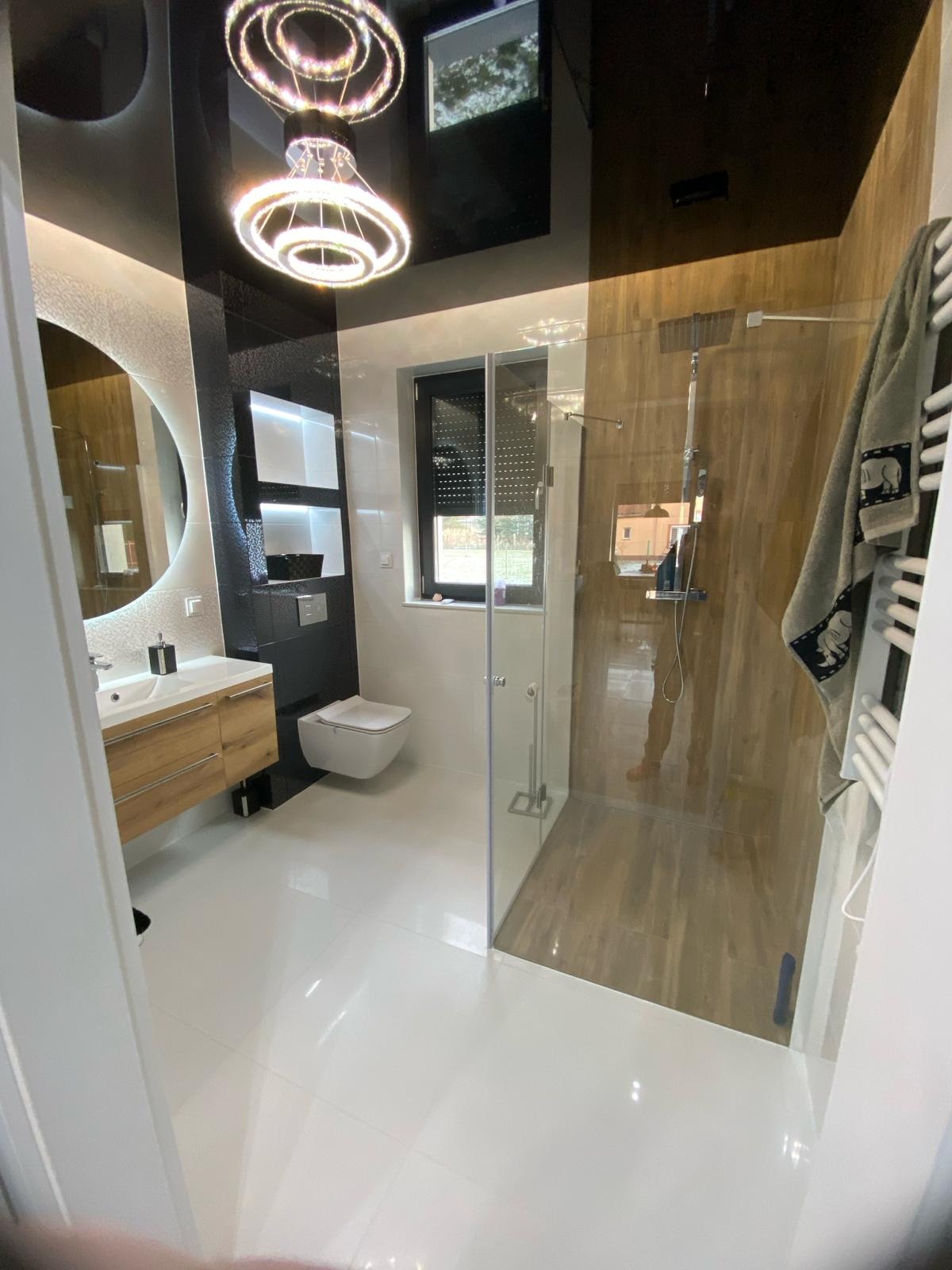 Nowoczesna łazienka z kabiną prysznicową typu walk-in, drewnianymi akcentami i okrągłym lustrem z podświetleniem LED. Białe płytki podłogowe i designerska lampa sufitowa.