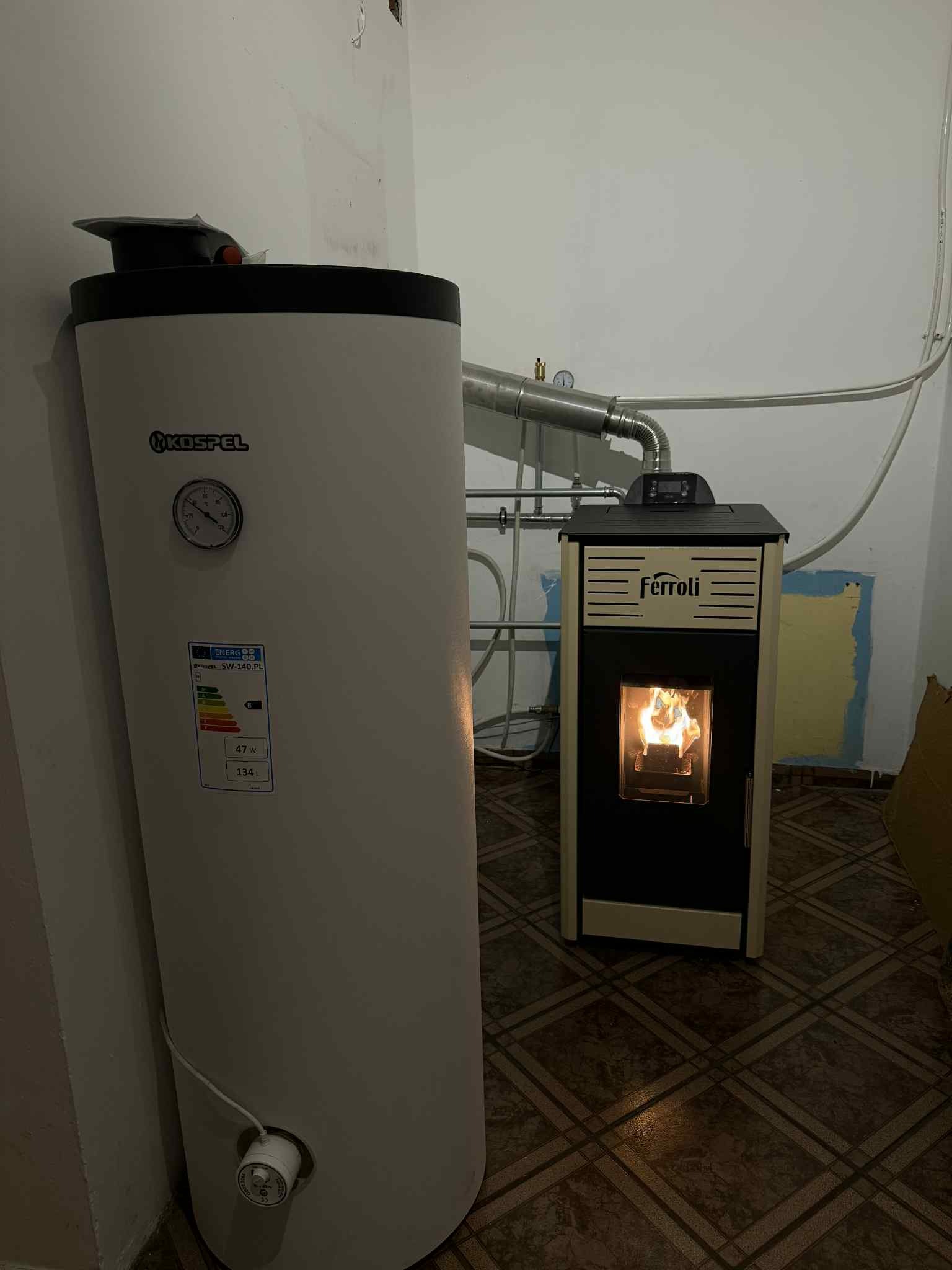 Biały bojler Kospel z widocznym wskaźnikiem i etykietą energetyczną, obok piec Ferroli z płomieniem w palenisku, połączone rurą. Widoczne instalacje.