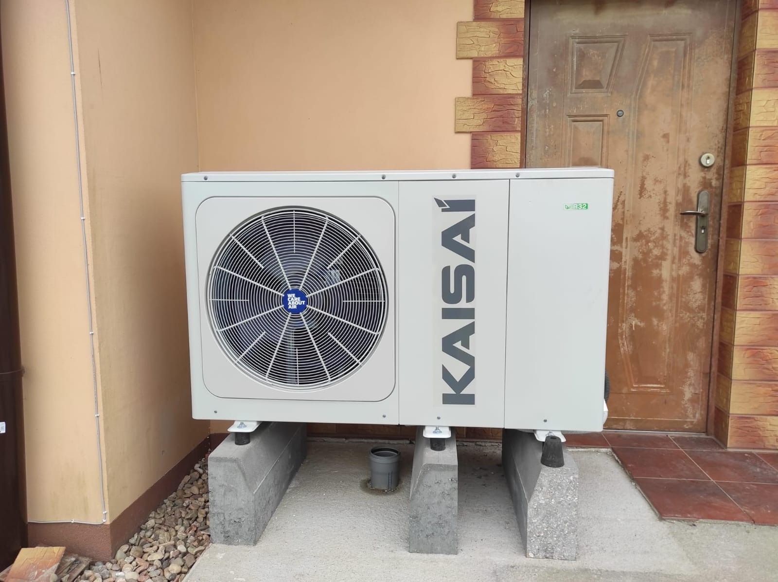 Biała pompa ciepła Kaisai, zamontowana na betonowych podstawach przed beżową ścianą i brązowymi drzwiami wejściowymi. Widoczny fragment rynny i kamieniste podłoże.