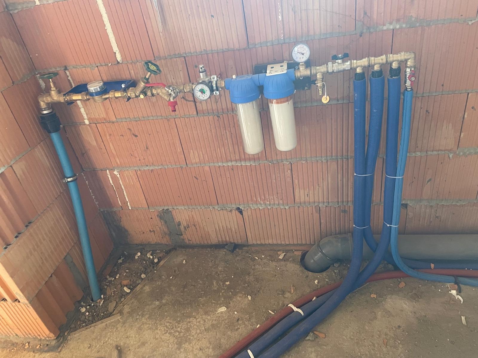 Instalacja wodna z filtrami i zaworami, widoczna na tle ceglanej ściany w surowym stanie. Rury w izolacji prowadzone wzdłuż ściany i podłoża.