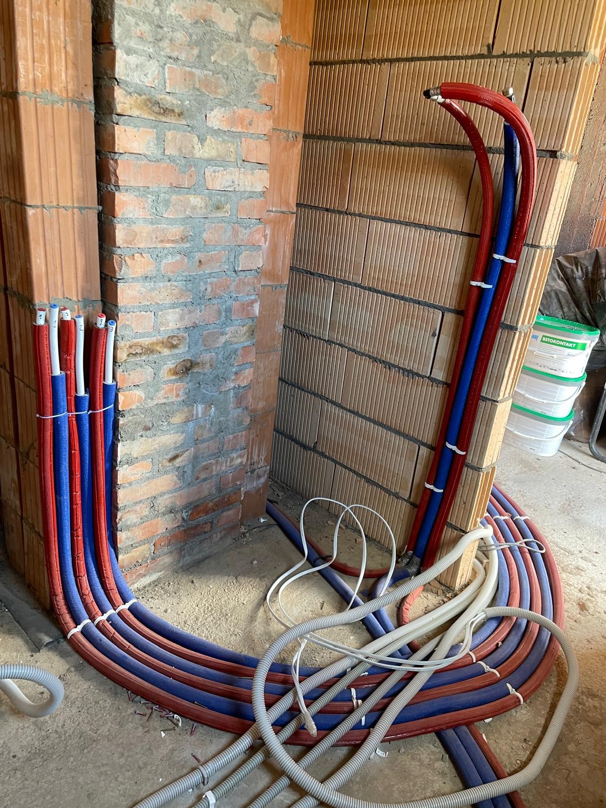 Instalacja ogrzewania podłogowego w trakcie montażu, rury czerwono-niebieskie ułożone w pętle na surowej posadzce, widoczne fragmenty ścian z cegły dziurawki.