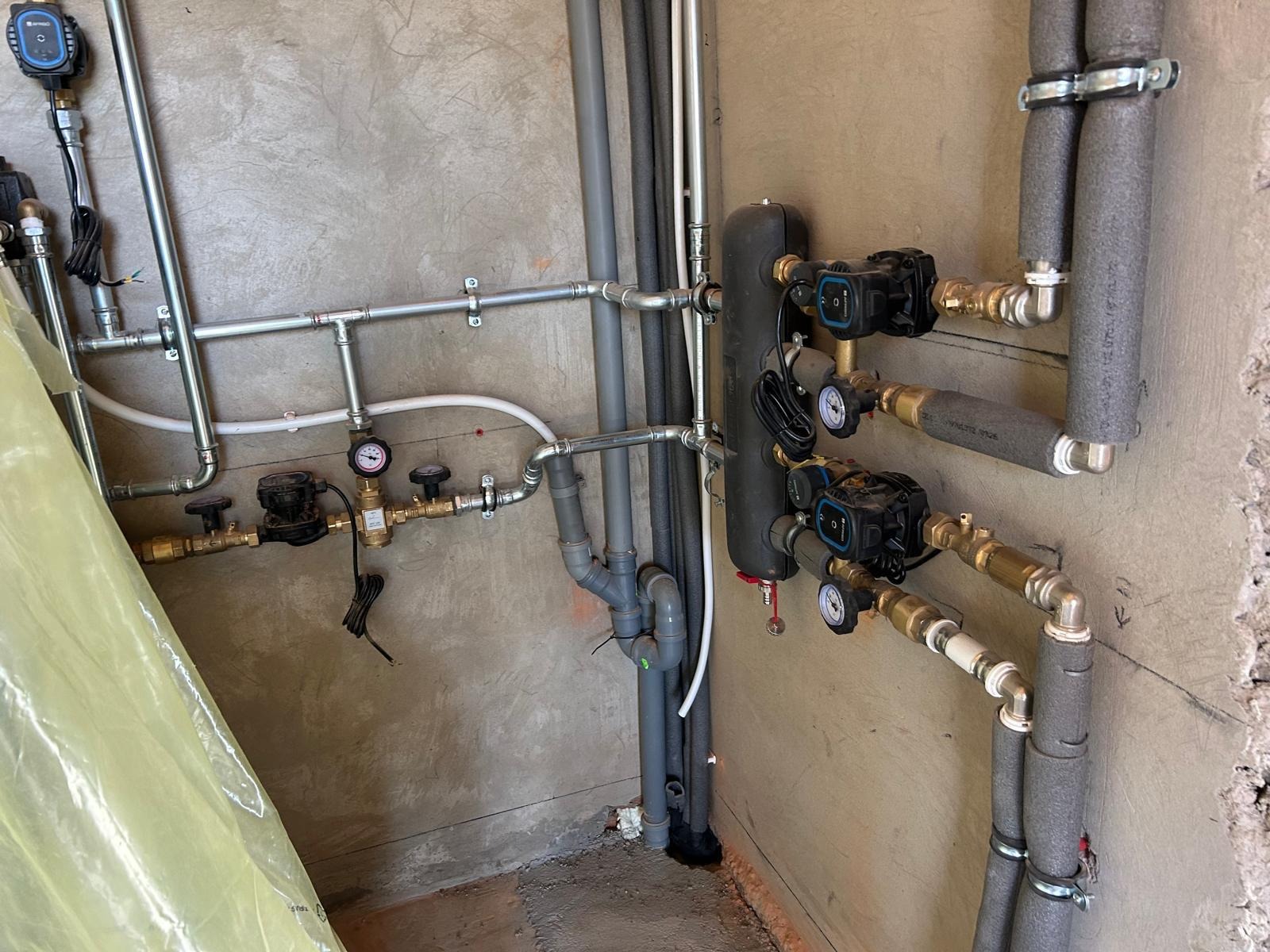 Złożona instalacja grzewcza z rurami, pompami obiegowymi i zaworami, zamontowana na surowej ścianie. Widoczne elementy izolacji termicznej na rurach.