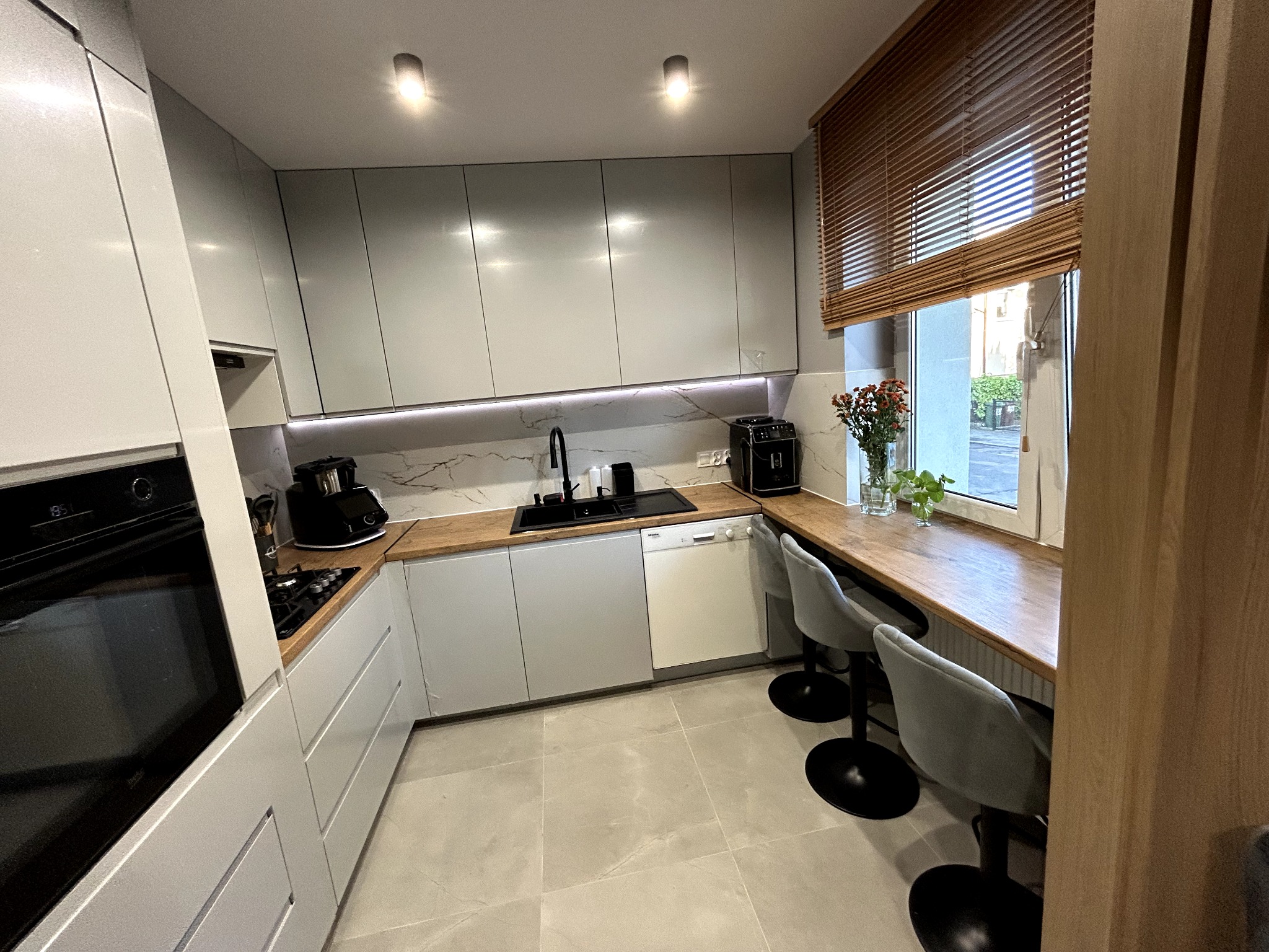 Nowoczesna kuchnia w szarościach z drewnianymi akcentami: blat, żaluzje i wyspa barowa przy oknie. Minimalistyczny design, oświetlenie LED i marmurowy backsplash.