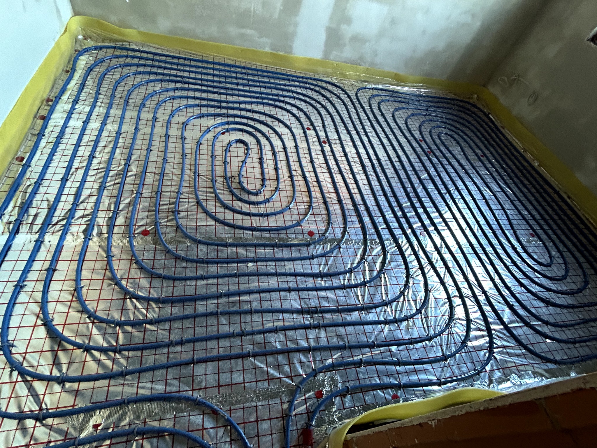 Niebieskie rury ogrzewania podłogowego ułożone spiralnie na folii izolacyjnej z czerwoną siatką, przygotowane do zalania posadzką w nowym budynku.