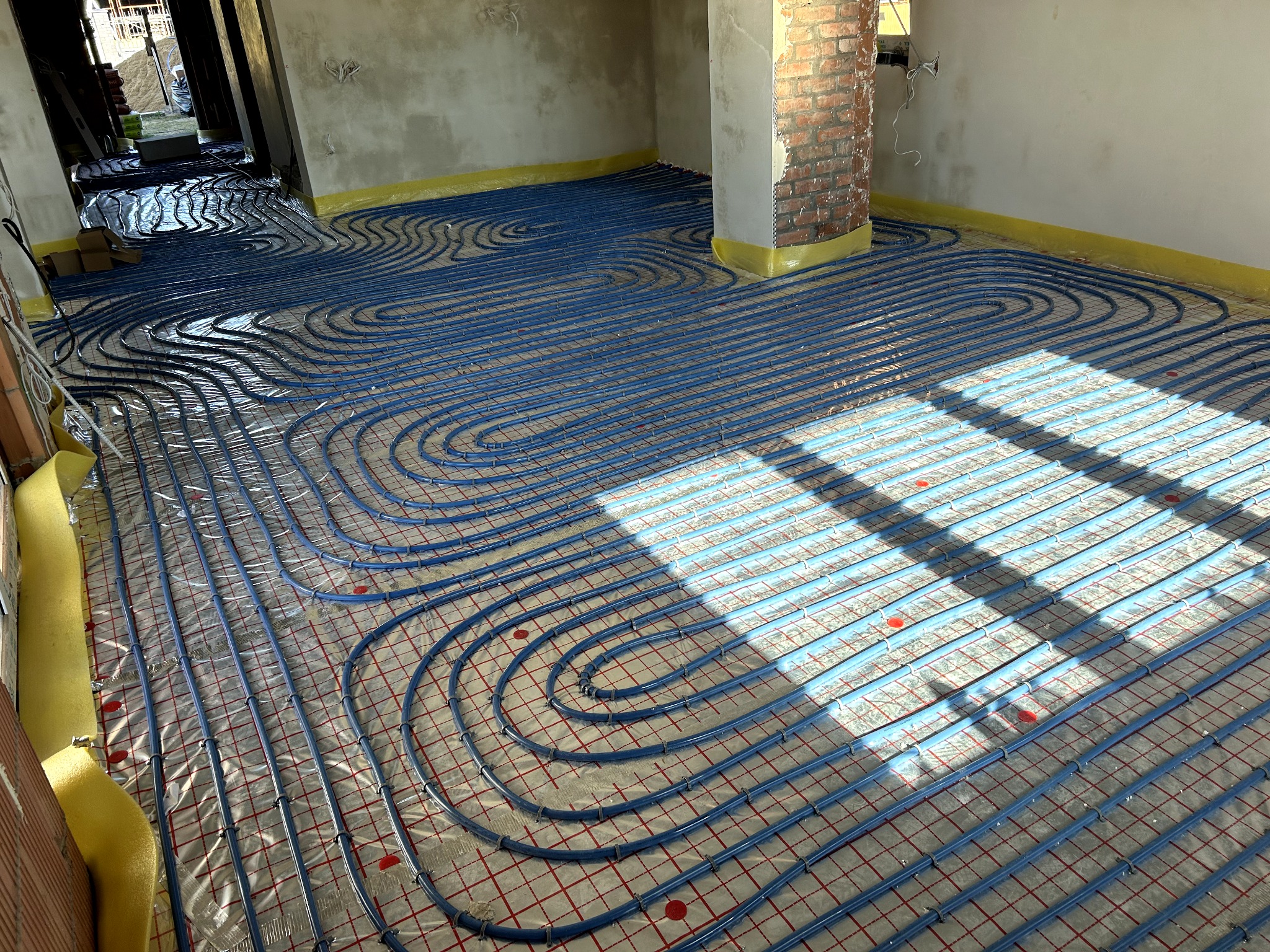 Niebieskie rury ogrzewania podłogowego ułożone na folii z czerwoną siatką, widoczne fragmenty ścian w surowym stanie i kolumna z cegły. Żółta taśma zabezpieczająca.
