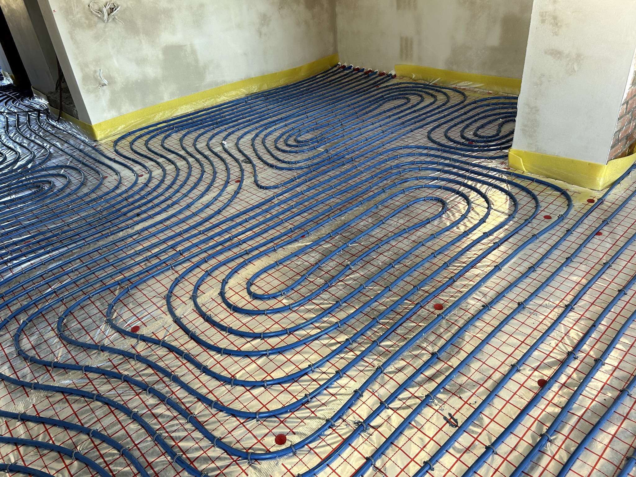 Niebieskie rury ogrzewania podłogowego ułożone na folii z czerwoną siatką, zabezpieczone żółtą folią przy ścianach w surowym wnętrzu. Widok z góry, ukośnie.