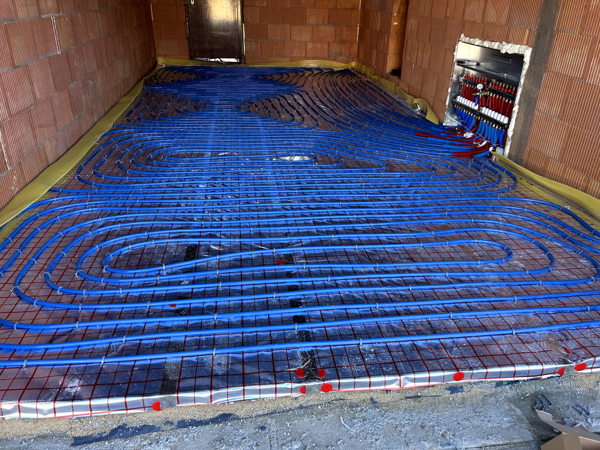 Niebieskie rury ogrzewania podłogowego ułożone spiralnie na folii z siatką, widoczne rozdzielacze w ścianie z cegły. Żółta taśma brzegowa.