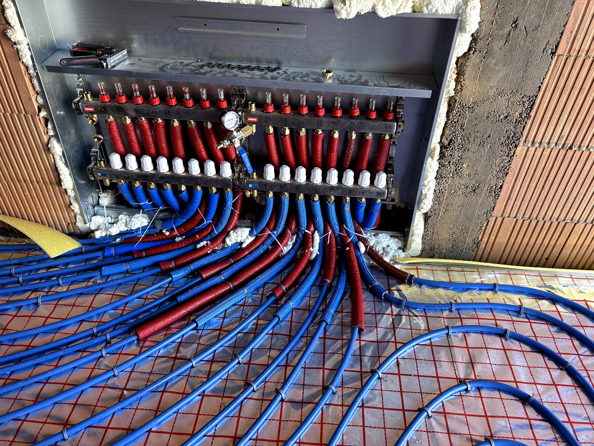 Rozdzielacz ogrzewania podłogowego z rurami w kolorze niebieskim i czerwonym, zamontowany na ścianie z cegły. Widoczne narzędzia i izolacja piankowa.