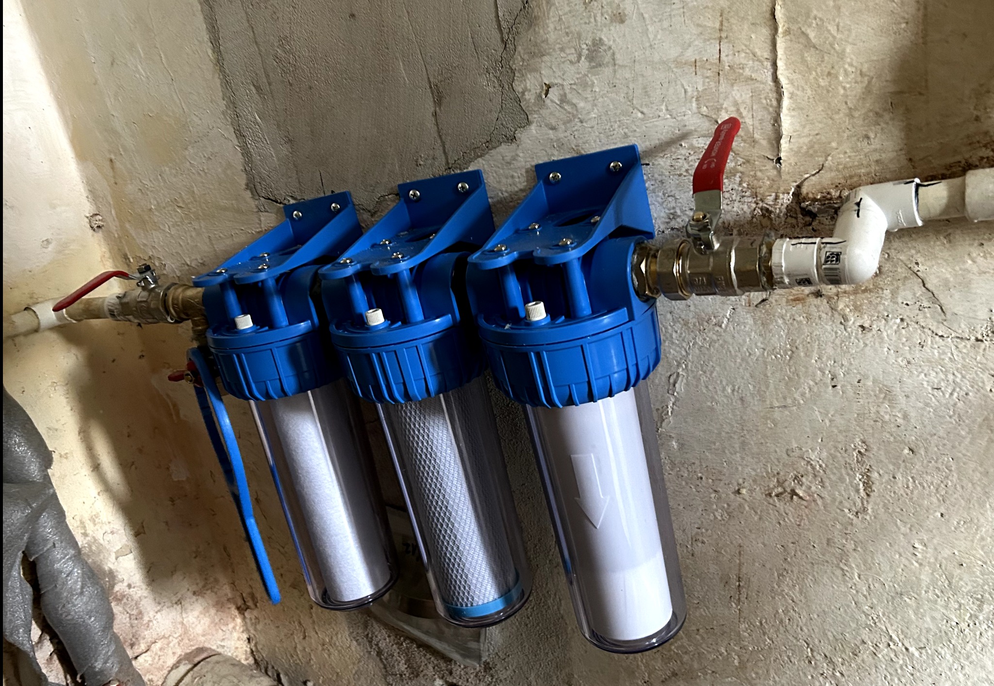 Potrójny filtr wody z niebieskimi obudowami zamontowany na ścianie z widocznymi rurami i zaworami. Filtracja wody w instalacji domowej.