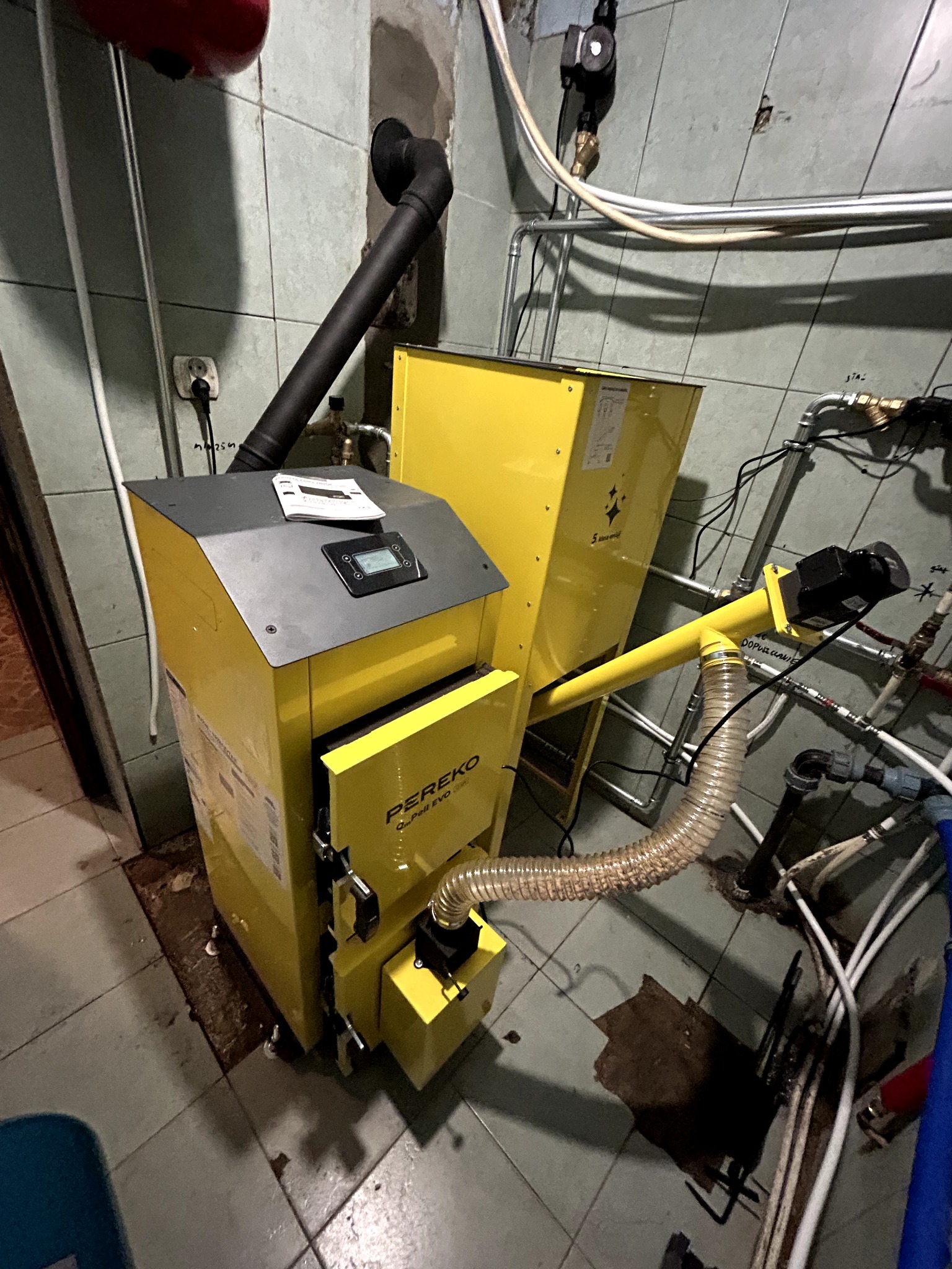 Żółty piec na pellet Pereko z czarnym kominem i podajnikiem, zainstalowany w kotłowni z widocznymi rurami instalacji CO i CWU na tle ściany z płytek.