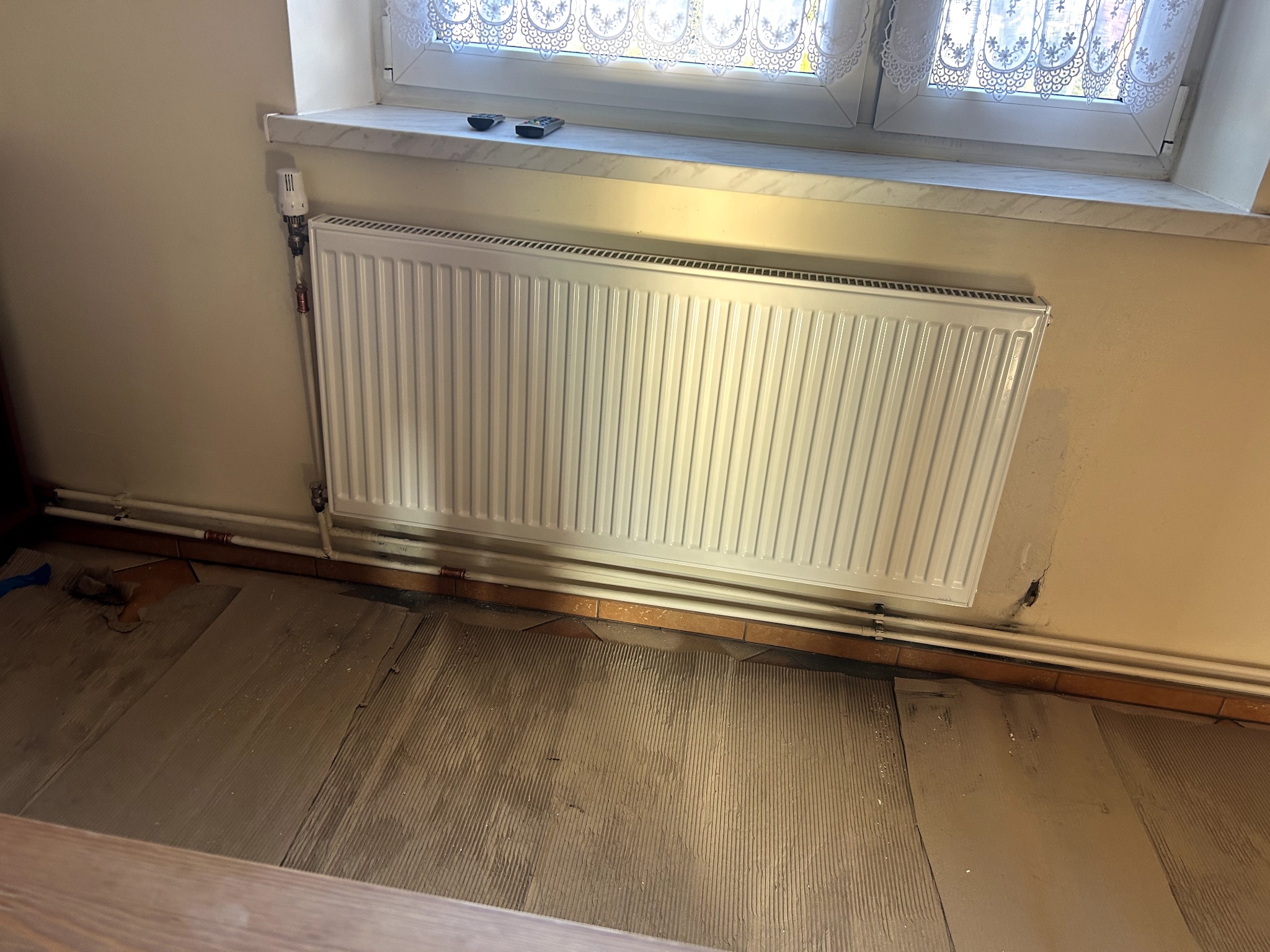 Biały grzejnik panelowy zamontowany pod oknem na beżowej ścianie. Widoczne rury doprowadzające wodę i termostat. Podłoga w trakcie remontu, z widocznymi śladami po starej posadzce.