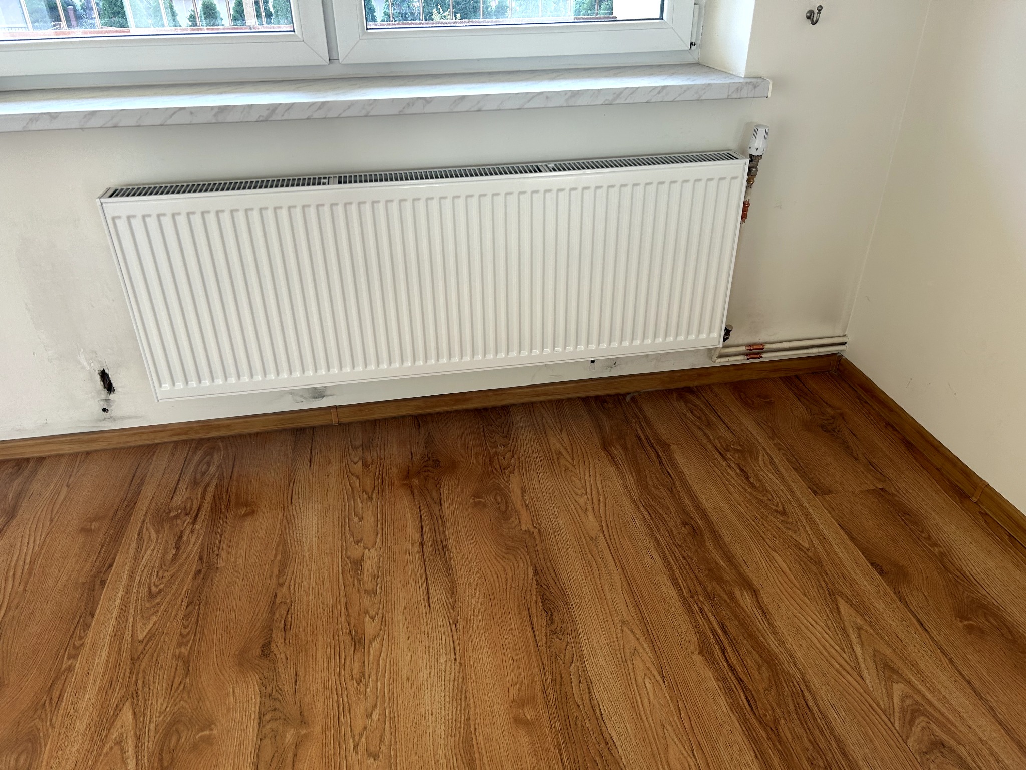 Biały grzejnik panelowy zamontowany pod oknem na ścianie z widocznymi śladami po instalacji. Na podłodze panele imitujące drewno. Widoczne rury doprowadzające.