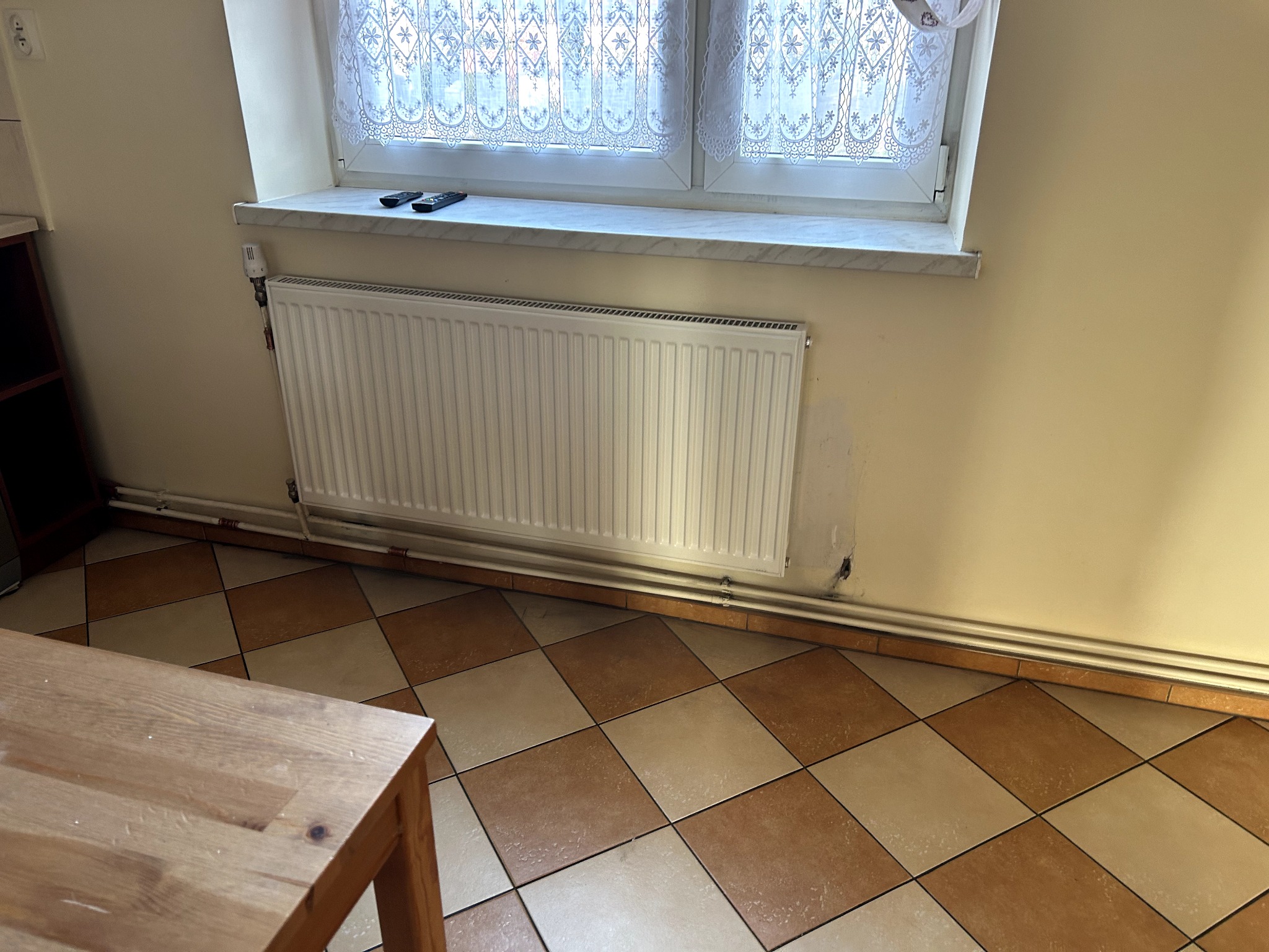 Biały grzejnik panelowy pod oknem z firanką w pokoju z brązowo-beżową podłogą w kratkę. Na parapecie leżą dwa piloty. Widoczne rury instalacji grzewczej.