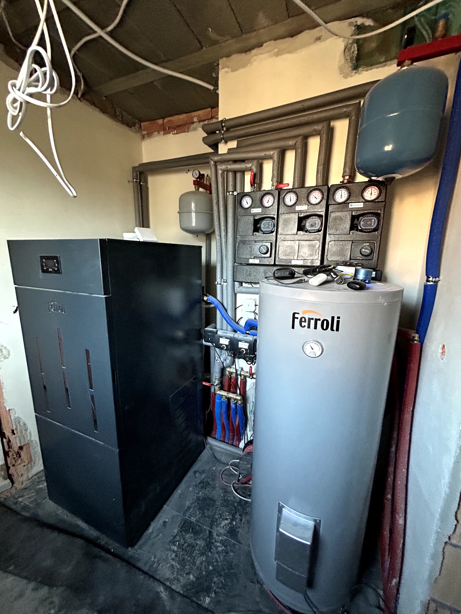 Czarny piec CO i szary zasobnik Ferroli w kotłowni. Widoczne rury, manometry i instalacja. Nierówne ściany i sufit. Ujęcie z dołu, podkreślające wysokość urządzeń.