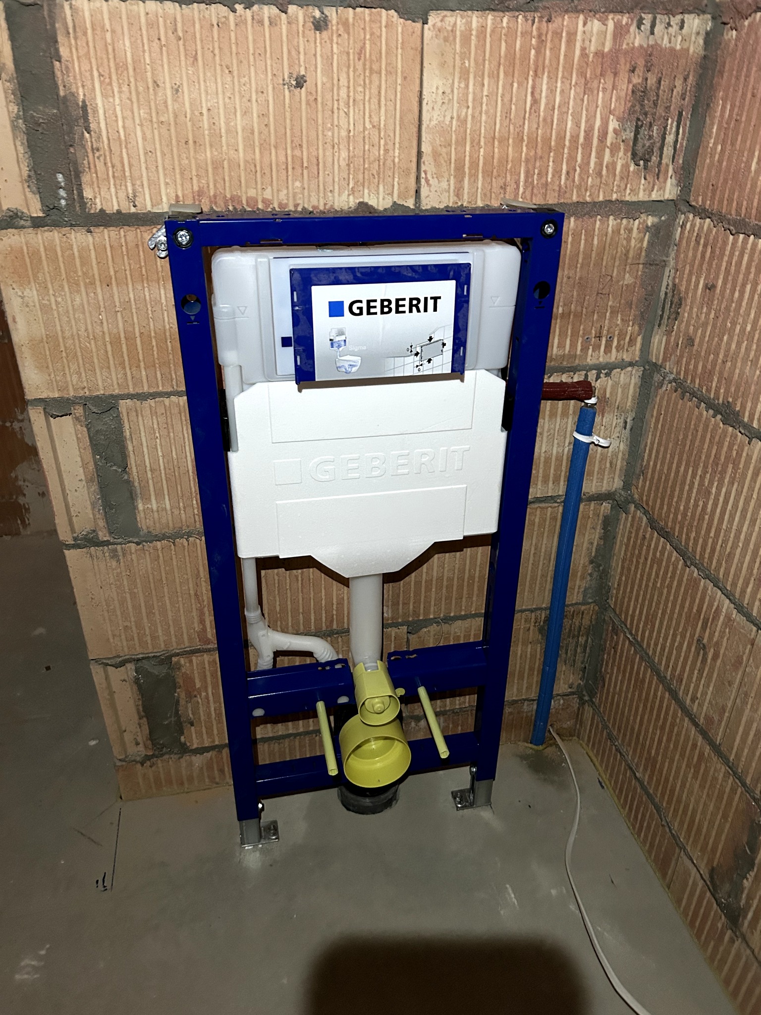 Stelaż podtynkowy Geberit zamontowany przy ceglanej ścianie, widoczne elementy instalacji wodnej i kanalizacyjnej. Etap montażu przed zabudową.