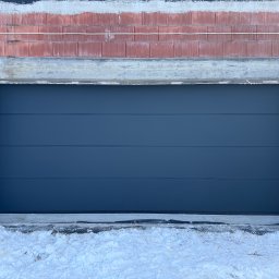 Andrzej Frąś montażownia - Zamknięta, segmentowa brama garażowa w kolorze antracytowym, zamontowana w surowym otworze budynku w trakcie budowy. Widoczne bloczki i ośnieżone podłoże.