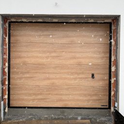 Andrzej Frąś montażownia - Garażowa brama segmentowa Hormann z drewnopodobnym panelem, czarną ramą i widocznymi elementami montażowymi w surowym otworze budynku, podczas opadów śniegu.