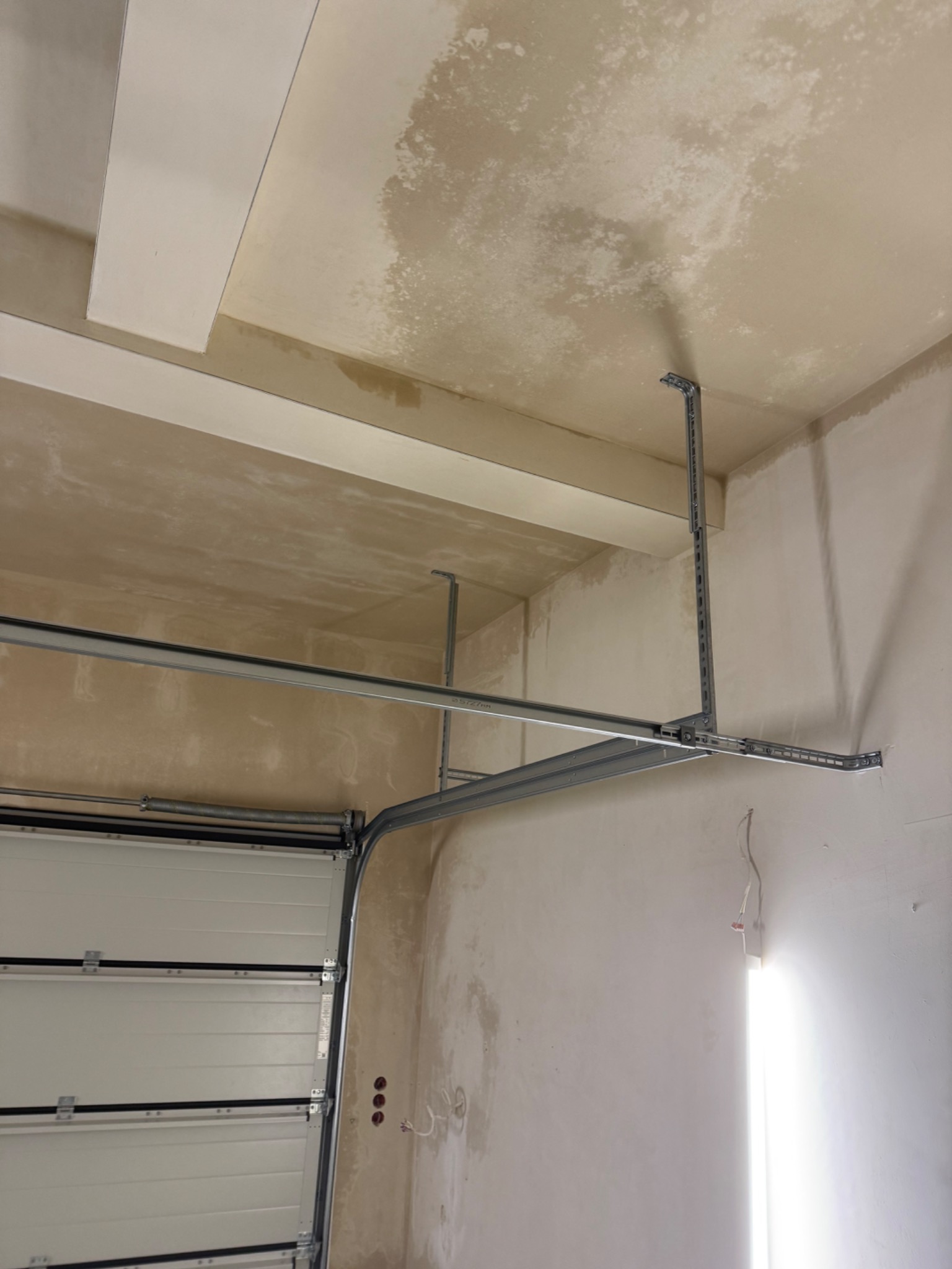 Wnętrze garażu w trakcie montażu bramy segmentowej. Widoczne prowadnice i elementy konstrukcyjne bramy, surowy tynk na ścianach i suficie, oświetlenie robocze.