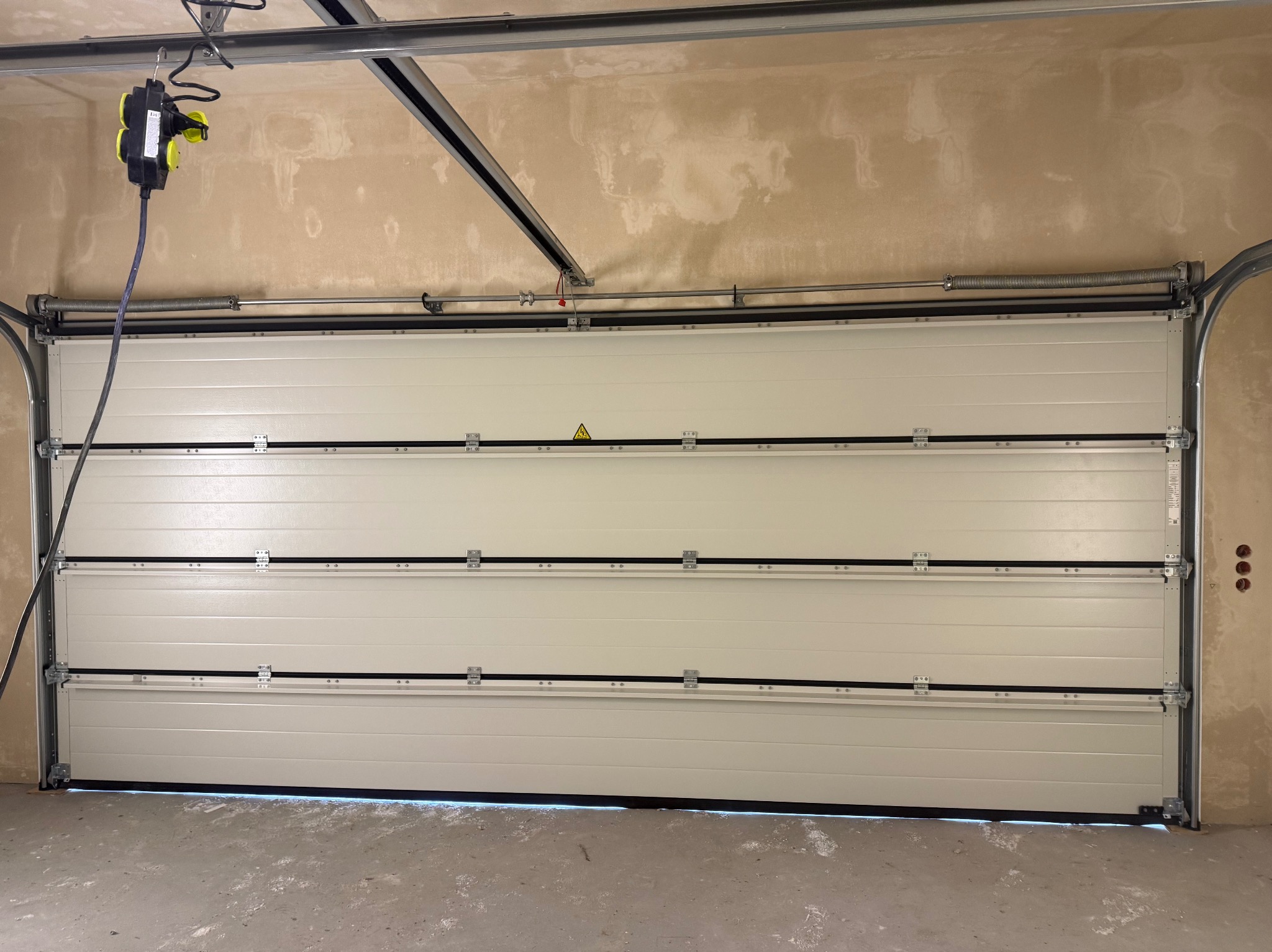Zamknięta, segmentowa brama garażowa w jasnym odcieniu, widoczna od wewnątrz garażu. Nad bramą widoczny mechanizm sprężynowy i silnik z podwieszonym przedłużaczem.