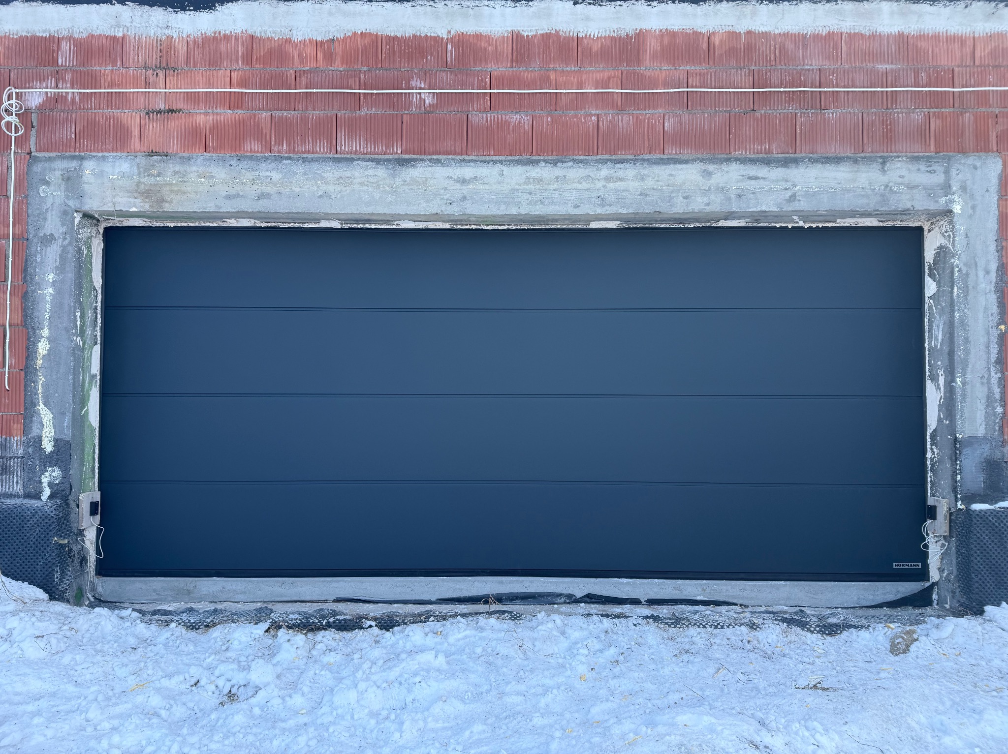Zamknięta, segmentowa brama garażowa w kolorze antracytowym, zamontowana w surowym otworze budynku w trakcie budowy. Widoczne bloczki i ośnieżone podłoże.