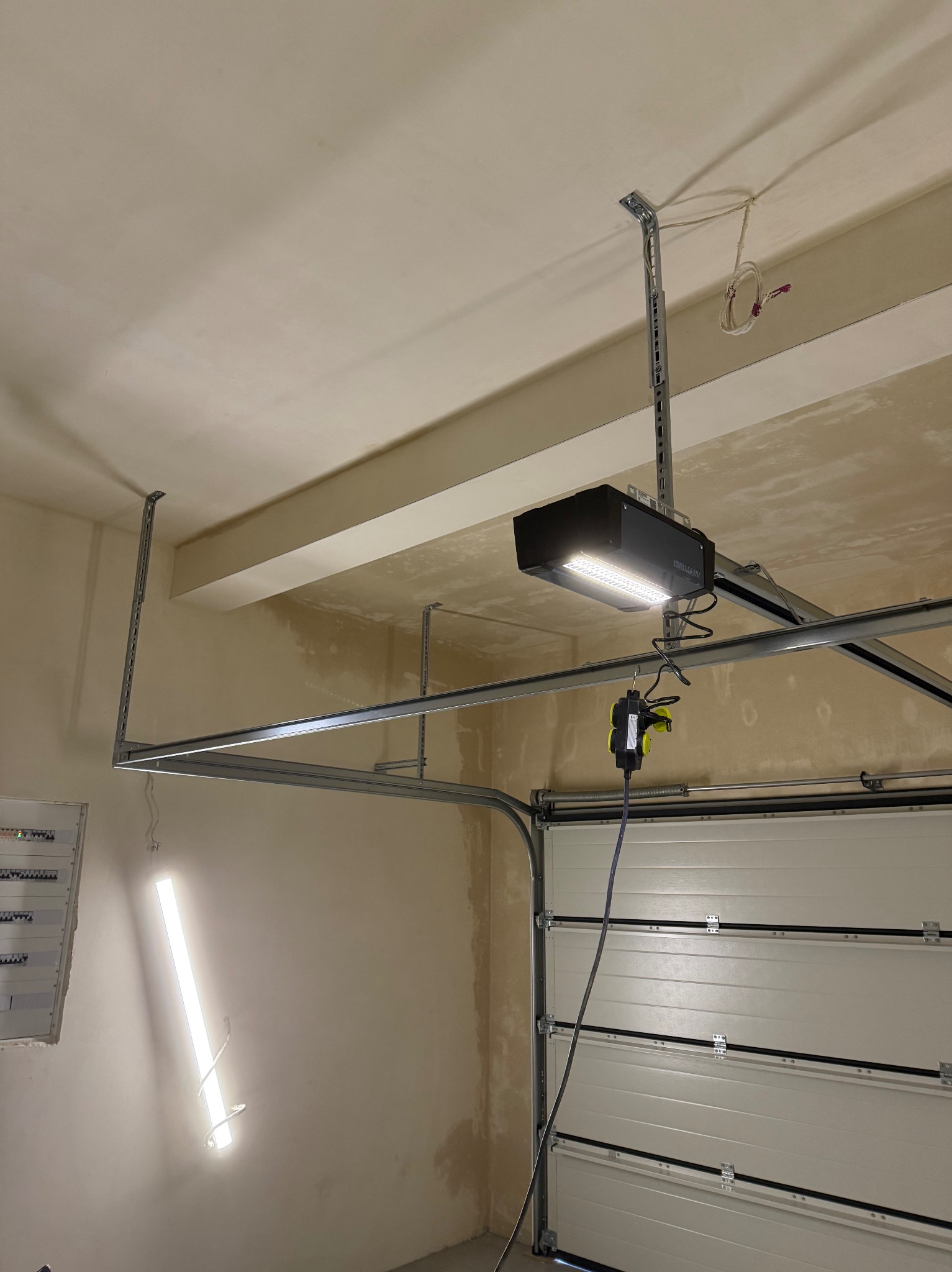 Zamontowany napęd bramy garażowej z oświetleniem LED, widoczne szyny prowadzące i podłączenie elektryczne. Ściany garażu w trakcie wykończenia.
