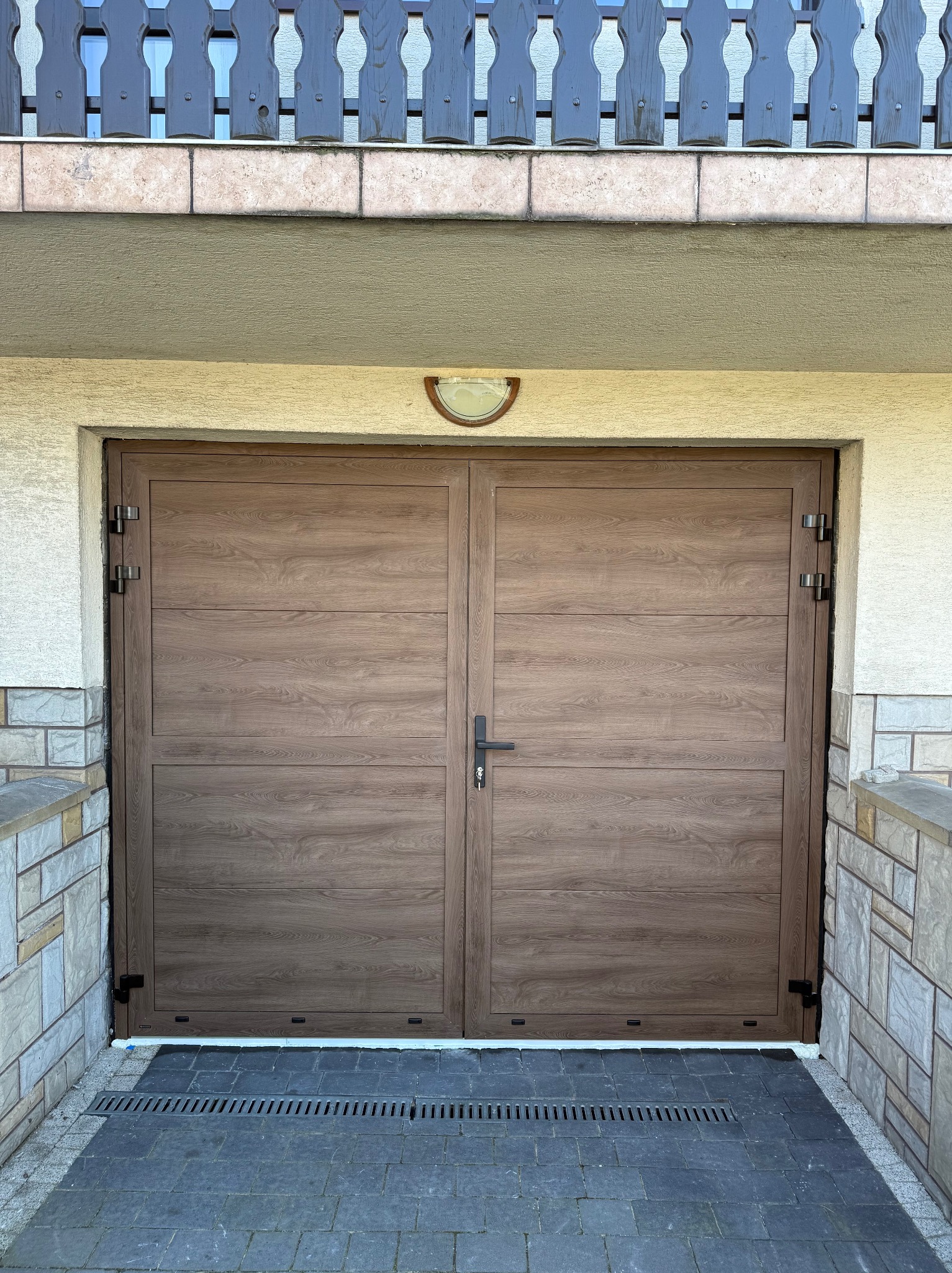 Dwuskrzydłowa brama garażowa w odcieniach brązu z poziomymi panelami, zamontowana w jasnym budynku z elementami kamiennymi i balustradą na górze.