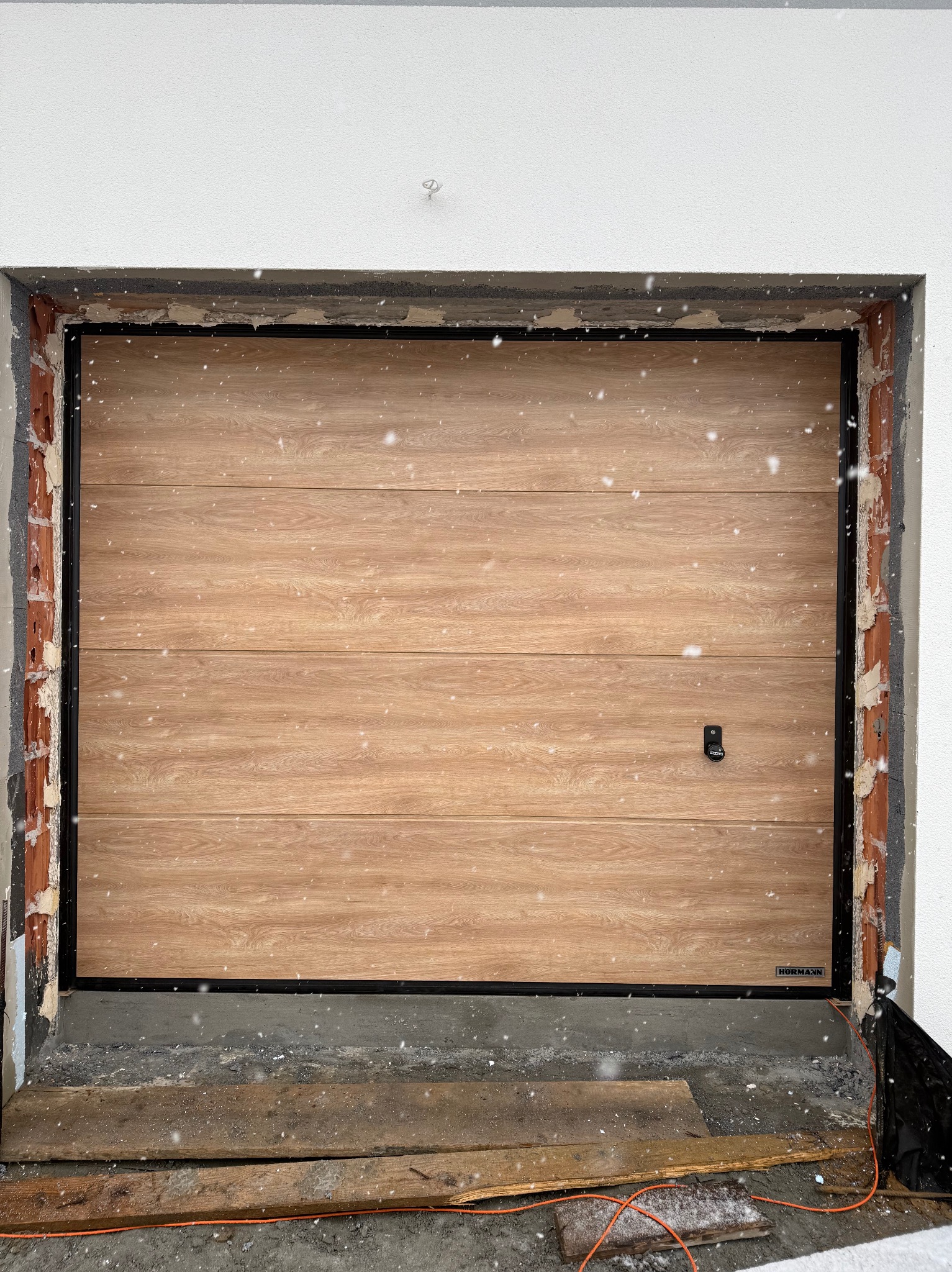 Garażowa brama segmentowa Hormann z drewnopodobnym panelem, czarną ramą i widocznymi elementami montażowymi w surowym otworze budynku, podczas opadów śniegu.