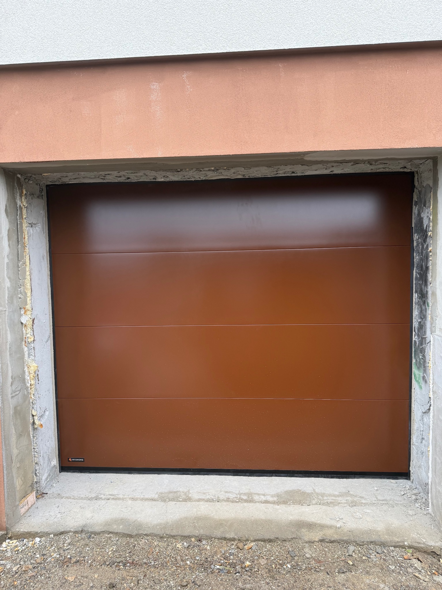 Brązowa brama garażowa segmentowa z poziomymi przetłoczeniami, widoczna od strony zewnętrznej, z logo producenta w lewym dolnym rogu. Surowy beton wokół otworu.