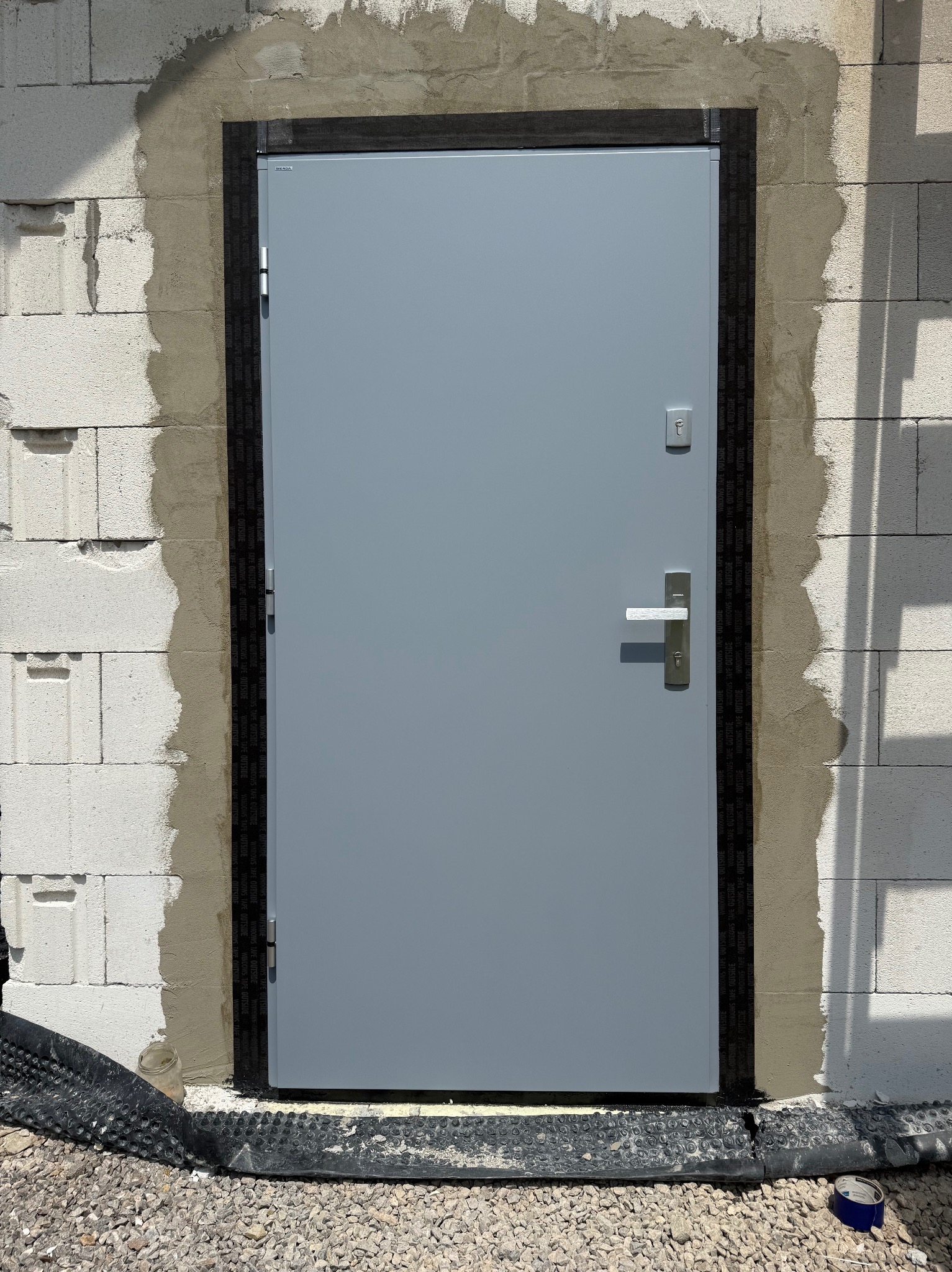 Szare drzwi zewnętrzne z ościeżnicą, zamontowane w surowym murze z betonu komórkowego. Widoczna taśma uszczelniająca i obróbka murarska wokół ościeżnicy.