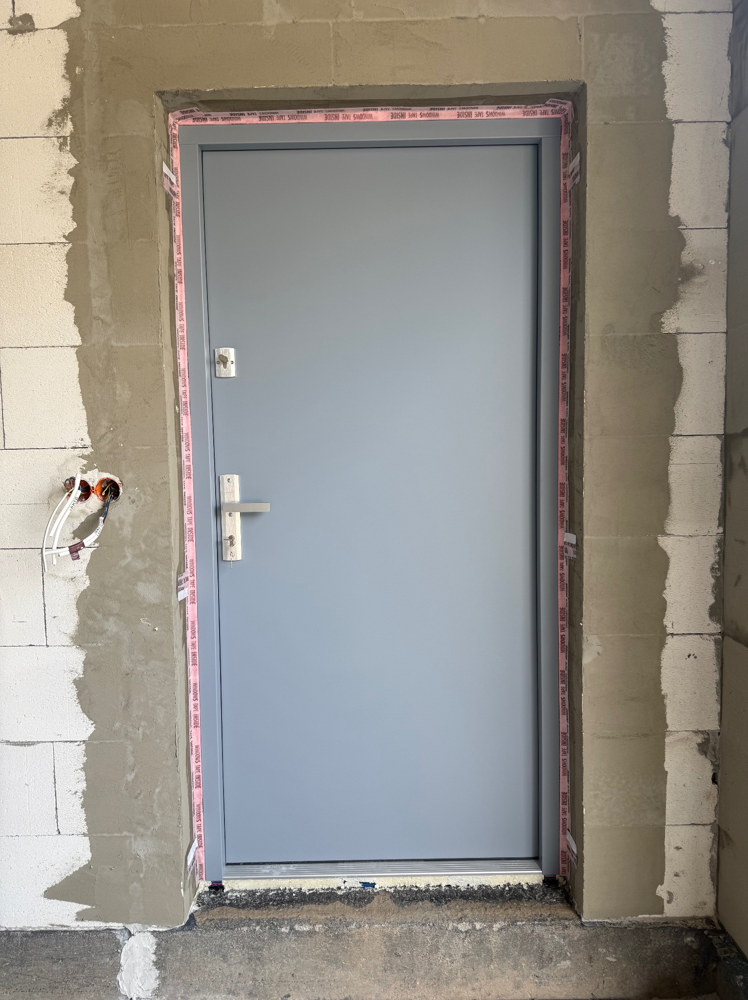 Szare drzwi wejściowe, zabezpieczone taśmą uszczelniającą, osadzone w surowym murze z widocznymi instalacjami elektrycznymi. Detal montażu.