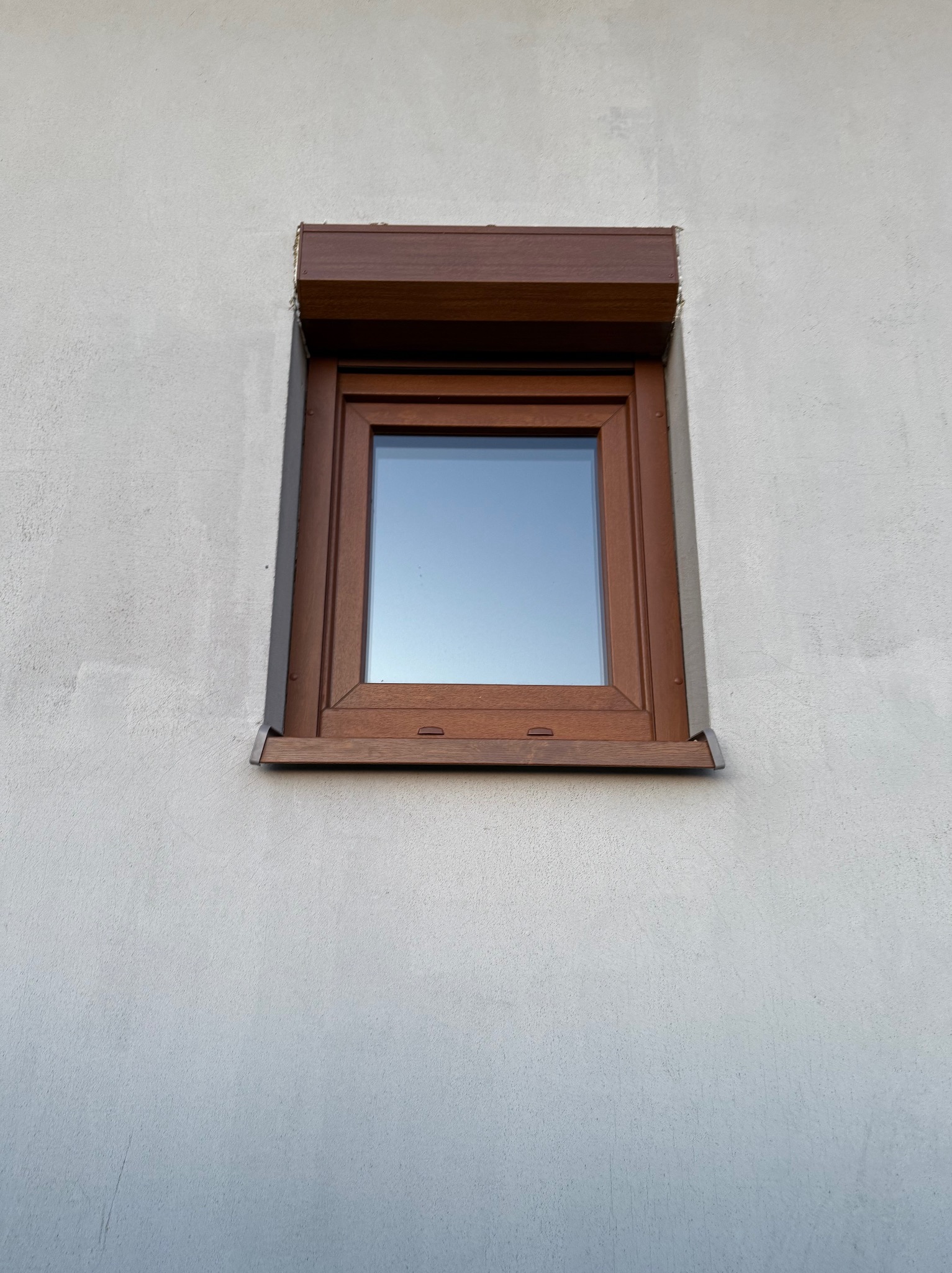 Okno z brązową roletą zewnętrzną na szarej elewacji. Widoczne detale montażu i faktura tynku. Ujęcie z dołu, podkreślające solidność wykonania.