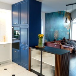 TOMASZEWSKI MEBLE PAWEŁ TOMASZEWSKI - Niebieska zabudowa kuchenna z piekarnikiem i szafką z ryflowanymi szybami, na tle salonu z dekoracyjną ścianą i sofą z dziećmi. Nowoczesne wnętrze.